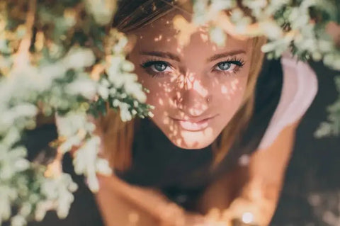 Smiling woman framed by flowers with sunlight on her face, symbolizing radiant skin glow