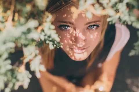 Smiling woman framed by flowers with sunlight on her face, symbolizing radiant skin glow