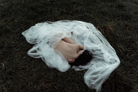 Person lying on grass wrapped in sheer white fabric, symbolizing contrast of synthetic vs natural